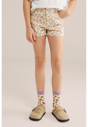 Copil purtând pantaloni scurți bej cu imprimeu leopard, șosete asortate cu margine violet și pantofi slip-on bej, stând în picioare pe un fundal simplu.