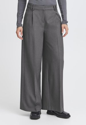 Person, der weite graue Hosen, schwarze Schuhe und ein langärmliges graues Oberteil trägt, steht vor einem schlichten hellen Hintergrund.
