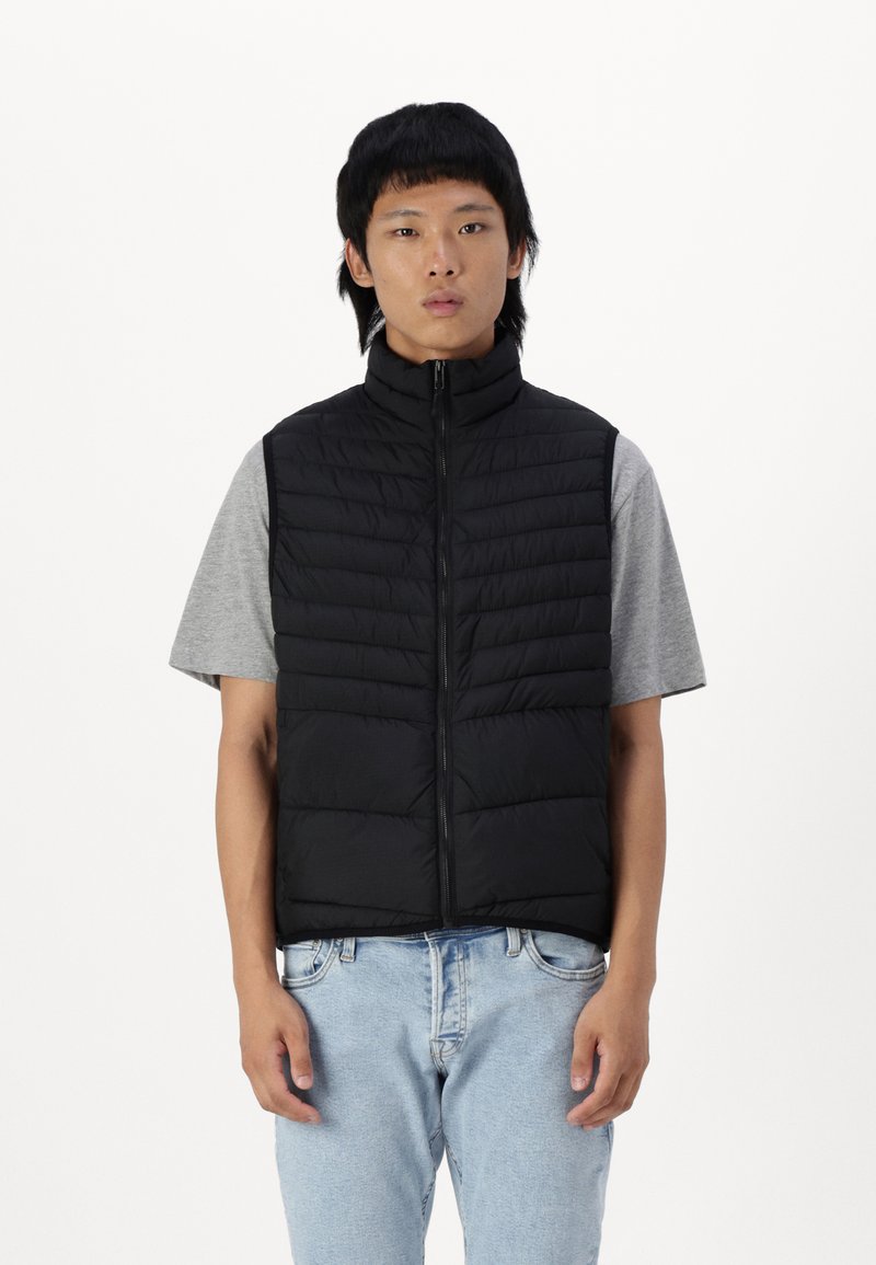 Zwarte gewatteerde vest met hoge kraag, ritsluiting, horizontale stiksels, gedragen over een grijze T-shirt. Casual fit, lichtgewicht stof, voorzakken.