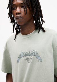 Tricou din bumbac verde deschis, cu un print grafic al cuvântului "Metallica" în albastru, cu un efect uzat și fraza "Justice for All."