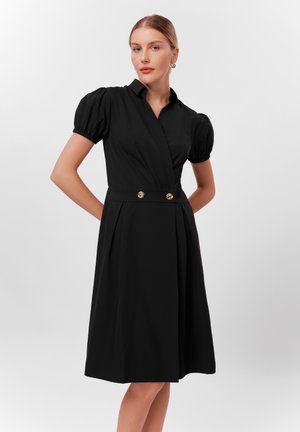 Robe noire avec un corsage ajusté, des manches bouffantes et un détail à la taille avec deux boutons dorés ; tissu lisse avec une jupe fluide.