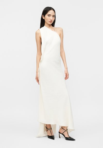 Pikkade tumedate juustega naine, kes kannab valget varrukateta asümmeetrilist kleiti ja musti kõrge kontsaga teravate otstega kingi, valgel taustal.