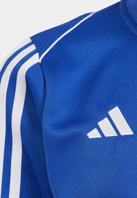 Blaues Sporttrikot aus strukturiertem Stoff, mit weißen Streifen an den Schultern und einem gestickten weißen Logo mit drei Streifen.