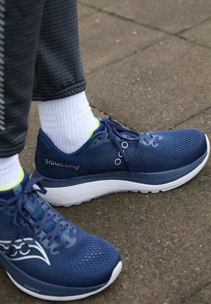 Blauwe netstof hardloopschoenen met witte zolen en grijze accenten, voorzien van een vetersluiting en een geperforeerde bovenkant voor ventilatie.