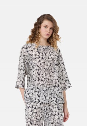 Donna con capelli ricci che indossa un top e pantaloni larghi abbinati con motivo floreale bianco e nero, in piedi con una mano in tasca.