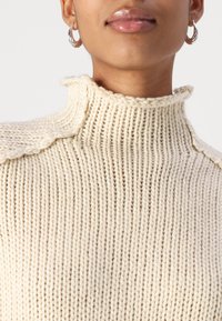 Suéter de punto marfil con un cuello alto, que presenta una textura acanalada y detalles laterales. El modelo lleva pendientes de aro plateados.