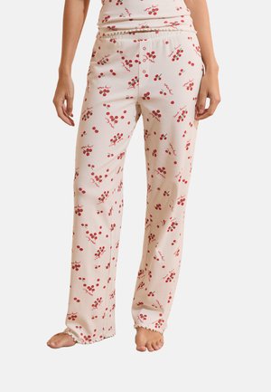Femme portant un pantalon de pyjama rose clair avec des motifs de cerises rouges et du texte rouge, debout pieds nus sur un fond blanc.