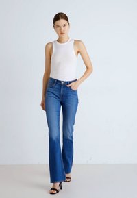 Biała top bez rękawów, niebieskie dżinsy w stylu dzwonów, slim fit, teksturowany dżins, wysoki stan, czółenka na kostkę, jednolite tło, modelka idąca.