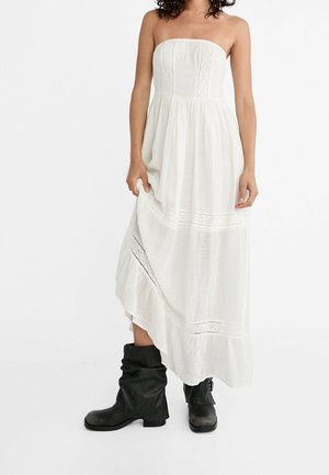 Robe maxi blanche sans bretelles avec détails en dentelle portée avec des bottes en cuir noir décontractées sur une personne debout, fond blanc.