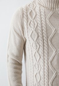 Cable-knit sweater in a light beige color. Features a high collar and intricate textured patterns on the front. Soft, thick fabric.