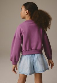 Felpa viola con maniche a palloncino e orlo a coste, abbinata a pantaloni corti in denim azzurro chiaro con bordo sfilacciato. Vista posteriore.