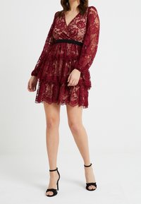 Abito in pizzo bordeaux con maniche lunghe, design a strati e una cintura nera. Presenta motivi floreali e un collo a V.