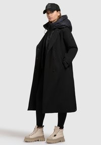 Manteau noir à double boutonnage avec une doublure à capuche volumineuse, associé à des bottes beiges montantes et à des leggings noirs. La casquette arbore un design de logo.