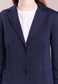 Blazer navy de tela suave, con cuello estilo notch, dos botones y bolsillos frontales. Textura suave y corte ajustado.
