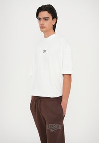 Camiseta de algodón blanca de manga corta con el logo negro de Reebok. Pantalones deportivos marrones con cinturilla elástica y texto gris de Reebok en el costado.