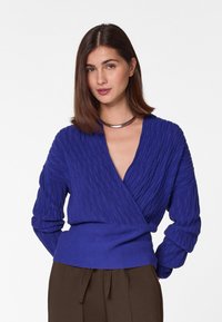 Lila gerippter Strickpullover mit tiefem V-Ausschnitt, langen Ärmeln und strukturiertem Stoff; kombiniert mit dunkelbraunen Hosen.