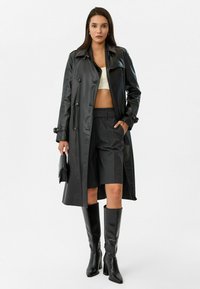 Zwarte leren trenchcoat met dubbele knopen, gecombineerd met grijze geplooide shorts en zwart knielaarzen. Eenvoudige zwarte handtas.