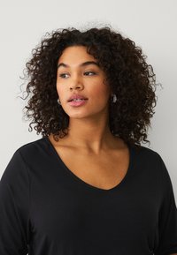 Maglietta nera a maniche lunghe con scollatura a V realizzata in materiale morbido, con una vestibilità comoda. Capelli ricci e orecchini ad anello sono visibili.
