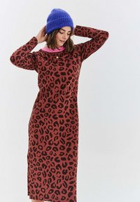 Žena upravující modrou pletenou čepici, oblečená v dlouhých hnědých šatech až po kotníky s černým leopardím potiskem a růžovým límcem.