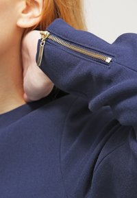 Námořnická mikina s přiléhavým střihem, obsahuje zlatý zip na rukávu a texturovanou látku. Detailní záběr na rukáv a zip.
