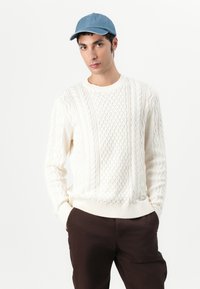 Cremefarvet kabelstrik sweater med teksturerede mønstre, ribbede manchetter og kant, parret med en blå denimkasket. Model iført brune bukser.