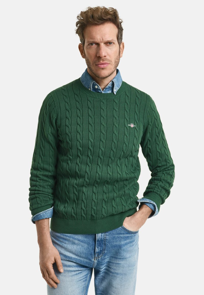 Grüner Kabelstrickpullover aus Wolle mit gerippten Bündchen und Saum, über einem Jeanshemd mit Kragen getragen, kombiniert mit blauen Jeans.