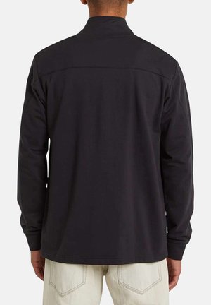 Homme portant une chemise noire à manches longues et un pantalon clair, vu de dos sur un fond uni.