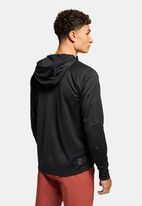 Schwarzes langärmliges Kapuzenshirt aus strukturiertem Stoff mit reflektierenden Akzenten und Daumenlöchern, kombiniert mit roten Shorts. Rückansicht dargestellt.