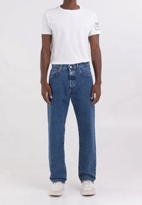 Jeans de mezclilla azul con un corte relajado, que cuentan con un diseño de cinco bolsillos, cierre frontal de botón y una ligera textura. Combinados con una camiseta blanca.