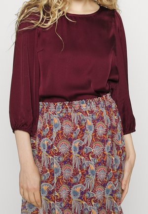 Kvinde iført en bordeaux langærmet bluse og et farverigt blomsterprintet skørt med elastisk talje, stående mod en ensfarvet baggrund.
