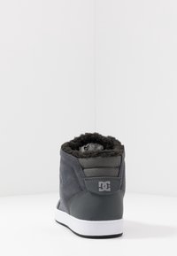 Harmaa mokkanahkainen nilkkuri, jossa on teksturoitu harmaa paneeli, tekoturkislining ja valkoinen kumipohja. Takana on logo ja pehmustettu kaulus.