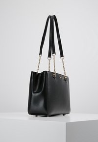 Sac en cuir noir avec des anses dorées en chaîne et des doubles poignées noires, posé sur un piédestal blanc contre un fond gris clair.