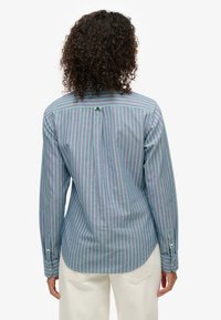 Femme aux cheveux bouclés portant une chemise à manches longues boutonnée à rayures verticales bleues et vertes, ainsi qu'un pantalon blanc, vue de dos.