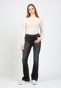 Hellbeige Rollkragenoberteil kombiniert mit hochgeschnittenen, schwarzen Schlagjeans, getragen mit weißen Sneakern, vor einem schlichten Hintergrund.