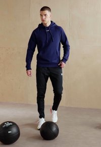 Marineblauwe hoodie met kangoeroezak, zwarte trainingsbroek en witte sneakers. Twee gewogen fitnessballen op de vloer. Minimale achtergrond.