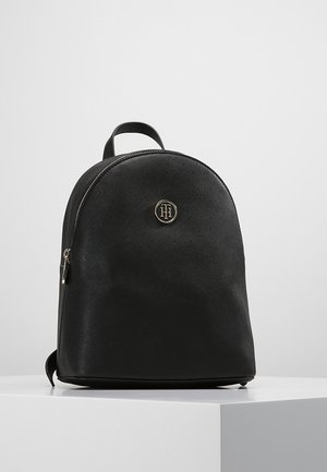 Sort textured rygsæk med afrundet top, enkelt håndtag, sølvlynlås og cirkulært sølvlogo på forsiden, stående på hvid overflade.