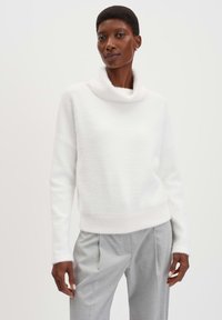 Pull à col roulé blanc, à la texture douce, avec une coupe décontractée et des bords côtelés, associé à un pantalon ajusté gris clair.