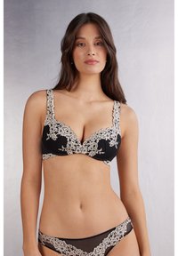 Soutien-gorge en dentelle noire avec broderie florale crème, larges bretelles et texture lisse. Inclus des culottes en dentelle noire assorties avec des accents floraux similaires.