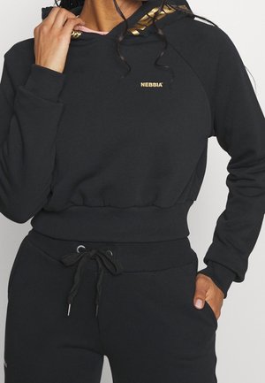 Vrouw draagt een zwarte cropped hoodie met goudkleurig "Nebbia" logo en bijpassende zwarte joggingbroek met trekkoord in de taille, hand in zak en houdt de capuchon kraag vast.