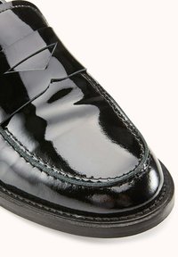 Mocassins en cuir verni noir avec une finition brillante, comportant un design cousu distinctif et une semelle plate et lisse.