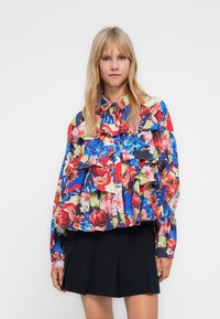 Blonde woman wearing a colorful floral ruffled blouse and a black pleated mini skirt, standing against a plain white background.