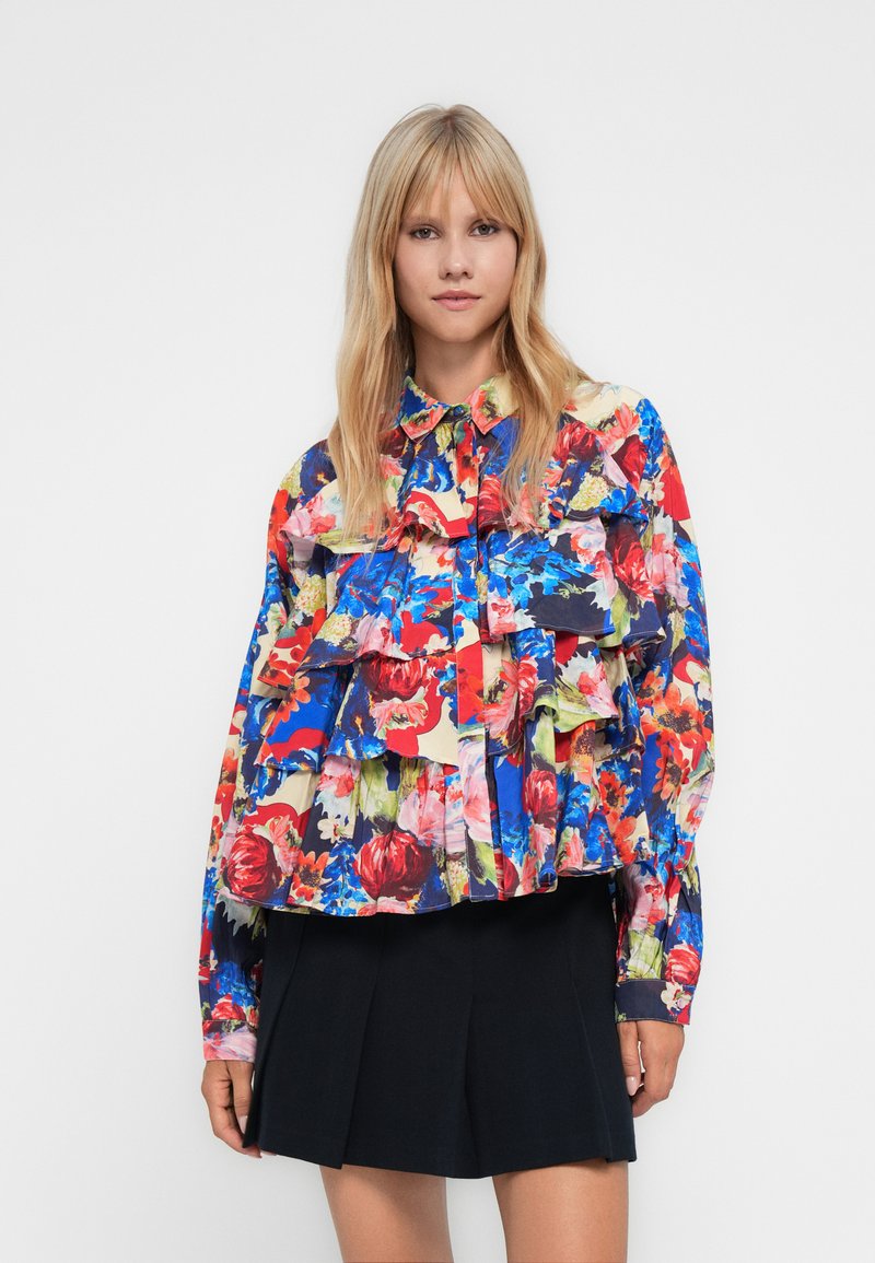 Blonde woman wearing a colorful floral ruffled blouse and a black pleated mini skirt, standing against a plain white background.