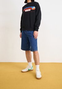 Zwarte trui met kleurrijke afbeelding en tekst, gecombineerd met blauwe denim shorts en witte sneakers met groene accenten. Effen achtergrond.