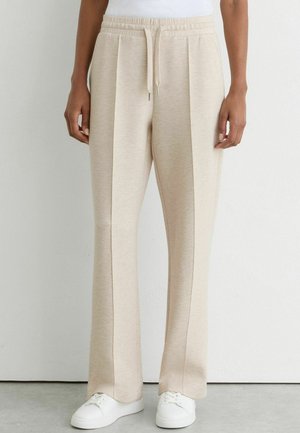 Person trägt beige weite Sweatpants mit vorderen Nähten und weiße Sneaker und steht an einer weißen Wand.