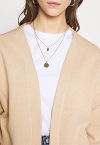 Cárdigan de punto beige con un ajuste holgado y abertura frontal, superpuesto a una camiseta blanca. Collares dorados de múltiples capas visibles.