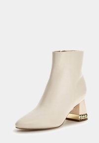 Stivale alla caviglia in pelle crema con punta appuntita e tacco a blocco geometricamente rifinito in oro, con un dettaglio a catena decorativo alla base.