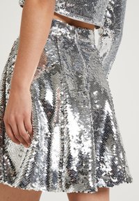 Falda de lentejuelas plateadas con un diseño acampanado, que presenta una textura suave y un cierre de cremallera oculto en la parte posterior. Las lentejuelas brillantes capturan la luz.