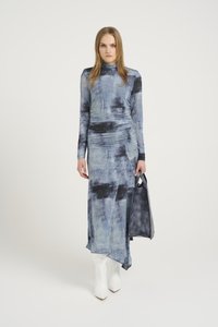 Vestido maxi em tie-dye azul com mangas longas, corpete ajustado e bainha assimétrica. Textura suave, combinado com uma mala escura e botas brancas até ao tornozelo.
