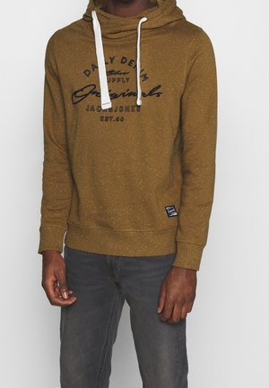 Mand iført en sennepsgul hættetrøje med hvide snore og sort tekst, kombineret med mørkegrå jeans, stående foran en ensfarvet baggrund.