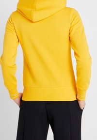Sweat à capuche jaune avec capuche, en tissu doux, doté de poignets et d'ourlet côtelés, avec un logo sur la manche. Porté avec un pantalon noir.